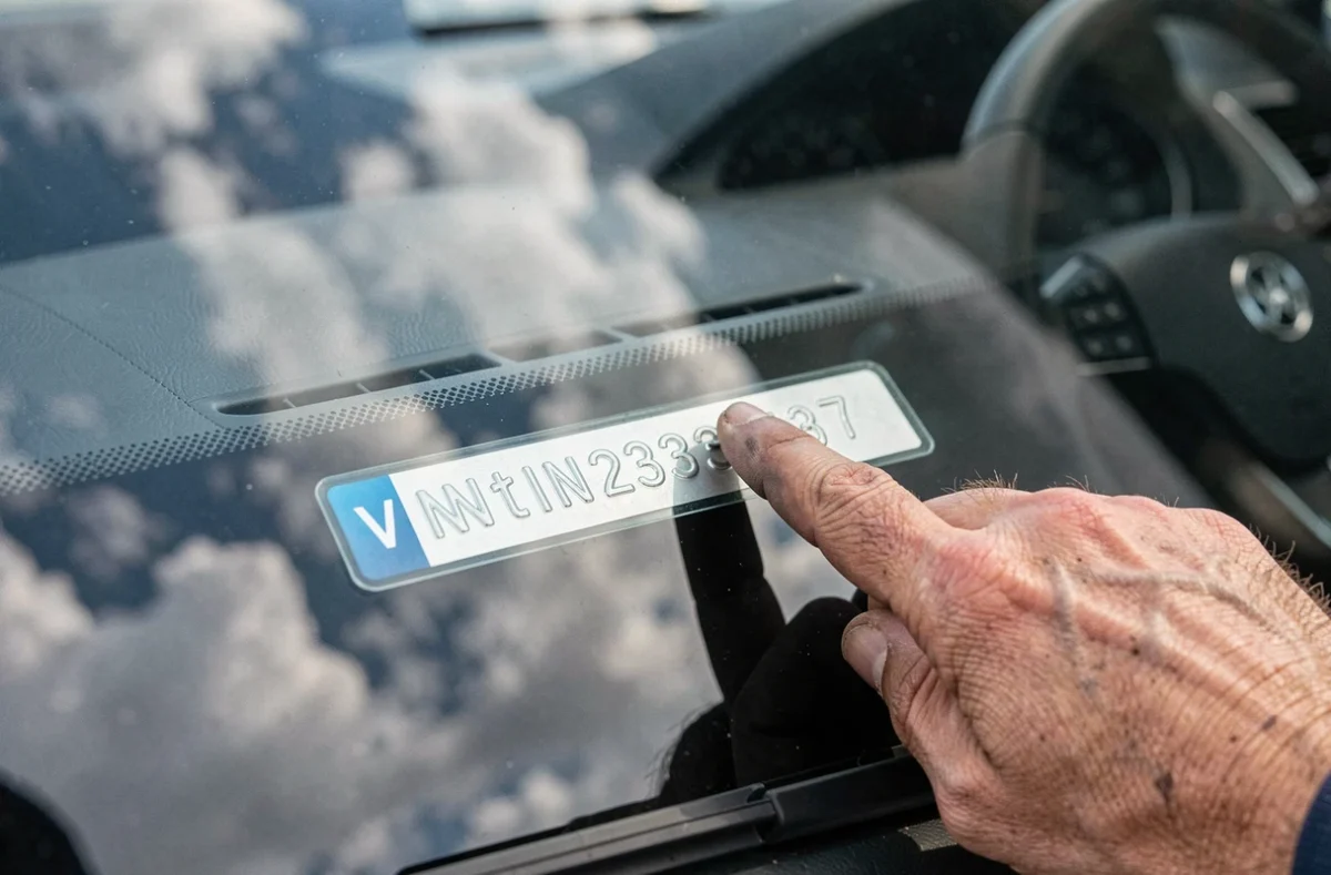Gros plan sur la plaque VIN au bas du pare-brise d'une voiture avec une main indiquant le numéro d'identification